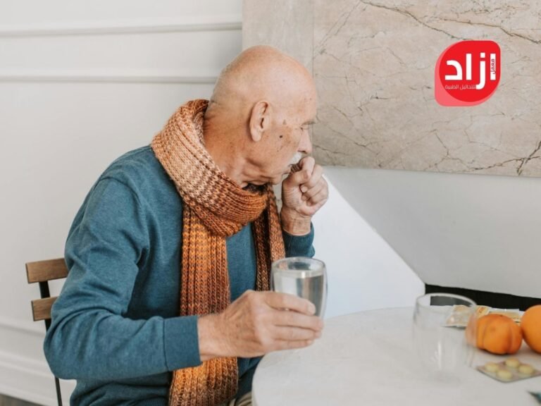 رجل مسن يرتدي وشاحًا ويجلس على طاولة أثناء إصابته بالكحة، ممسكًا بكوب ماء، في مشهد يعكس أعراض السعال المرتبطة بتغير الجو، مع شعار معمل ازاد للتحاليل الطبية في الخلفية.